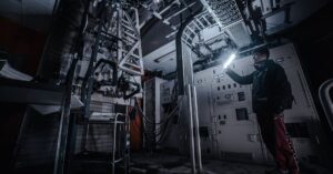 A technician using a flashlight to examine equipment in a dimly lit industrial control room.