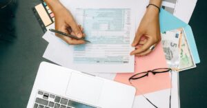 Hands writing on tax documents with laptop, glasses, and currency on desk.